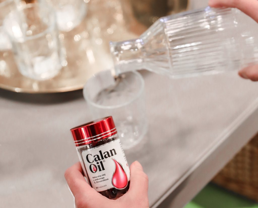opakowanie suplementu CalanOil na tle szklanki z wodą, nalewanej ze szkalnej butelki